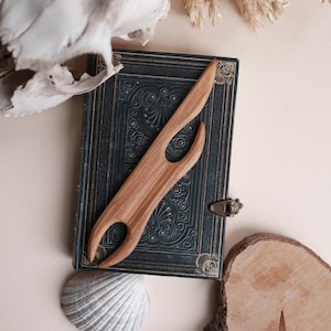 Op de afbeelding: Een houten garenklos ligt op een vintage blauw boek met gouden details. Een witte zeeschelp, een stuk hout en een schedel zijn rond het boek gerangschikt. De garenklos is lichtbruin en heeft een uniek, gebogen ontwerp.