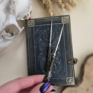 Puede incluir: Dos punzones plateados y bronceados descansan sobre un diario azul oscuro en relieve con un cierre decorativo. Una calavera y espigas de trigo secas están en el fondo. Los punzones tienen mangos ornamentados y puntas afiladas.