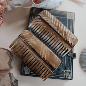May include: Two wooden combs with carved chevron patterns rest on a dark blue book with gold accents. The combs are light brown with white accents. A skull and dried flowers are in the background.
