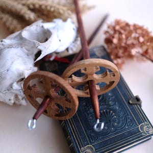 May include: Two wooden drop spindles with carved details, featuring a dark wooden shaft and a light-colored wooden wheel. The spindles rest on a dark blue book with gold accents, alongside a skull and dried flowers.