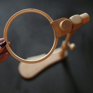 Puede incluir: Un soporte de aro de bordado de madera con un aro redondo y una base curva. El soporte es de madera clara y tiene un acabado natural. El aro se sujeta con una abrazadera de madera.