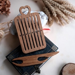 May include: Wooden weaving tools, including a rectangular loom with a heart-shaped handle and a shuttle, rest on a vintage book. The tools are made of light brown wood. A bundle of wheat and other decorative elements are in the background.