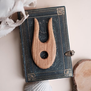 Puede incluir: Una herramienta de adivinación de madera marrón claro con un agujero circular, colocada sobre un libro antiguo azul oscuro con detalles dorados. La imagen incluye una concha, un cráneo y plantas secas, sugiriendo un tema místico.