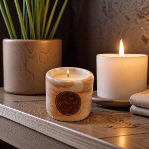 Pode incluir: Duas velas acesas, uma pequena com um design marmorizado pêssego e creme, e uma vela branca maior. Um vaso de madeira com vegetação está no fundo. A vela pequena tem um rótulo castanho com texto.