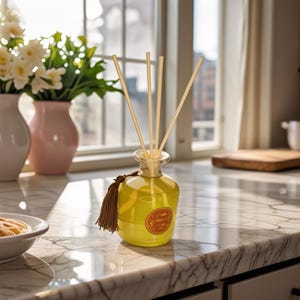 Könnte beinhalten: Eine gelbe Glas-Diffusorflasche mit Holzstäbchen und einer braunen Quaste. Die Flasche steht auf einer Marmorarbeitsplatte, neben einer weißen Schüssel und zwei Vasen mit Blumen. Das Etikett auf der Flasche ist orange.