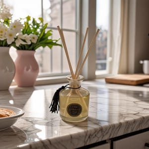 May include: A clear glass diffuser bottle with wooden reeds, filled with a light yellow liquid. A black tassel hangs from the neck. The bottle sits on a marble countertop, with a vase of white flowers in the background.