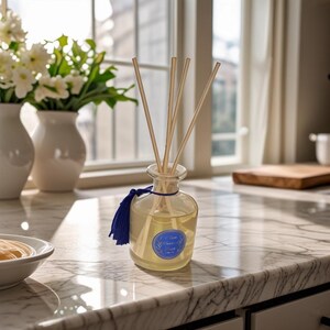 May include: A clear glass diffuser bottle with wooden reeds and a blue tassel. The bottle contains a yellow liquid and a blue label. White vases with flowers are in the background, creating a fresh, clean aesthetic.