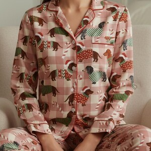 Może przedstawiać: Różowy zestaw piżamowy z powtarzającym się wzorem jamników w świątecznych swetrach i czapkach Świętego Mikołaja. Zestaw zawiera koszulę zapinaną na guziki z czerwonymi wykończeniami i pasujące spodnie. Tło ma jasno różowy i biały wzór w kratę.