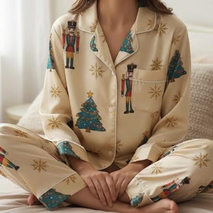 May include: Cream-colored pajama set featuring a repeating pattern of nutcrackers, Christmas trees, and snowflakes. The long-sleeved top and pants have teal and white trim. The pajamas are made of a soft, silky material.