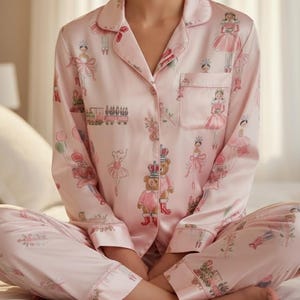 Peut inclure: Ensemble de pyjama rose clair avec un haut boutonné et un pantalon. L'ensemble présente un motif répété de ballerines, d'ours en peluche et de soldats de plomb. Le pyjama est en matière douce et soyeuse.