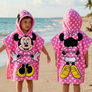 May include: Pink hooded beach towel with white polka dots and a Minnie Mouse graphic. The towel features a large Minnie Mouse character on the front and back, with yellow shoes and a bow. The towel is designed for children.