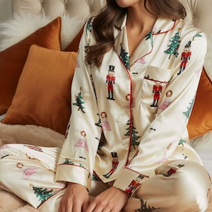 May include: A cream-colored pajama set featuring a repeating pattern of nutcrackers, Christmas trees, and ballerinas. The long-sleeved button-down top has a notched collar with red piping, a single chest pocket, and matching long pajama pants.