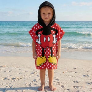 May include: A red and white polka-dot hooded beach towel featuring a Mickey Mouse design. The towel has a black hood and a large Mickey Mouse graphic on the front, with yellow shoes and white gloves. The child is standing on a sandy beach.