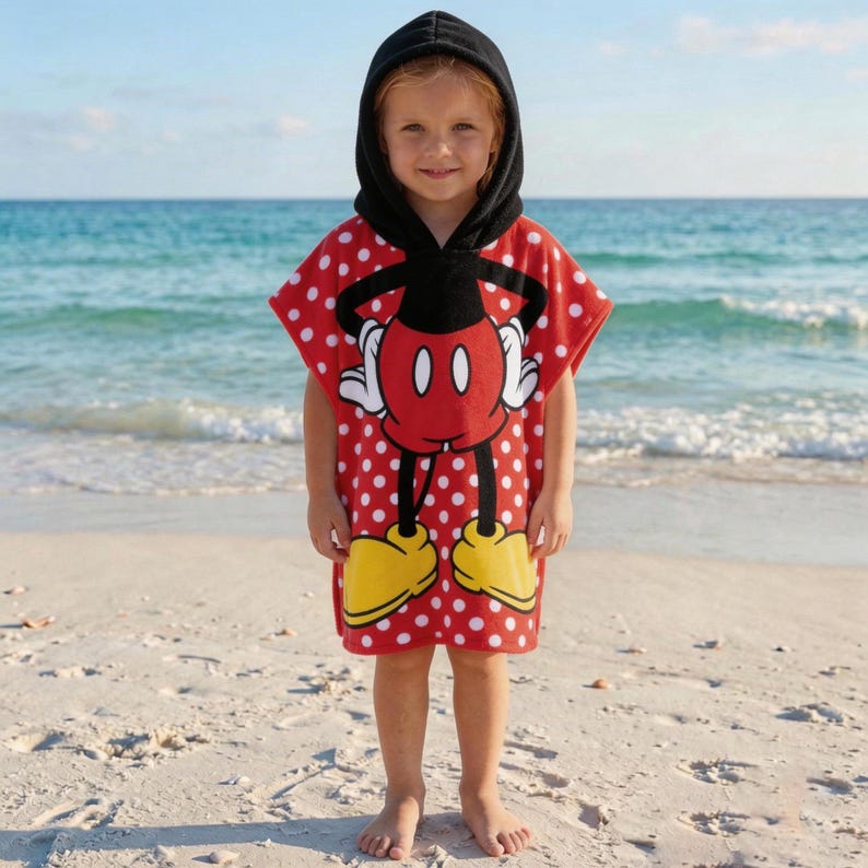 May include: A red and white polka-dot hooded beach towel featuring a Mickey Mouse design. The towel has a black hood and a large Mickey Mouse graphic on the front, with yellow shoes and white gloves. The child is standing on a sandy beach.