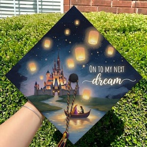 May include: A square graduation cap topper featuring a dark night sky with glowing floating lanterns, a large castle, and two figures in a small boat on water. The text reads ON TO MY NEXT dream in white lettering with a multi-colored tassel attached.