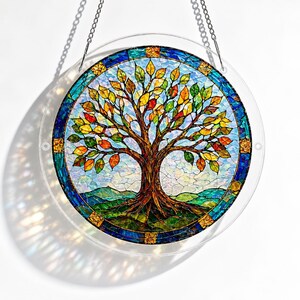 Pu&ograve; includere: Un acchiappasole circolare in vetro colorato raffigurante un albero con foglie multicolori nelle tonalit&agrave; del giallo, arancione e verde. L'albero &egrave; posto su uno sfondo di colline verdi e un cielo azzurro, racchiuso da un bordo decorativo blu.