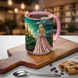 Puede incluir: Una taza de caf&eacute; con asa rosa con un dise&ntilde;o de vidriera que representa a una mujer con un vestido rosa, un castillo y un cielo turquesa. La taza est&aacute; llena de caf&eacute; y est&aacute; sobre una tabla de cortar de madera.