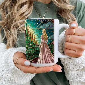 Puede incluir: Taza de cer&aacute;mica blanca con una ilustraci&oacute;n estilo vidriera. El dise&ntilde;o representa a una mujer con un vestido rosa mirando un castillo verde bajo un cielo estrellado. Los detalles dorados enmarcan la obra de arte, creando un tema caprichoso y de cuento de hadas.