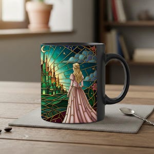 Puede incluir: Taza de cer&aacute;mica negra con un dise&ntilde;o de vidriera que representa a una mujer con un vestido rosa mirando un castillo verde. El dise&ntilde;o incluye una luna creciente, estrellas y nubes. La taza tiene un asa negra y est&aacute; sobre un mantel gris.