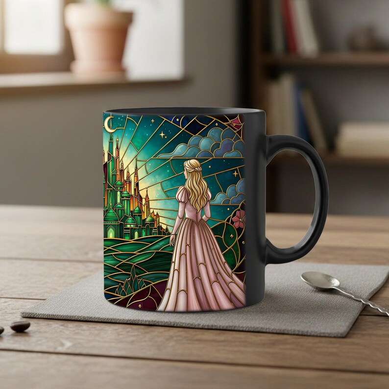 Puede incluir: Taza de cer&aacute;mica negra con un dise&ntilde;o de vidriera que representa a una mujer con un vestido rosa mirando un castillo verde. El dise&ntilde;o incluye una luna creciente, estrellas y nubes. La taza tiene un asa negra y est&aacute; sobre un mantel gris.