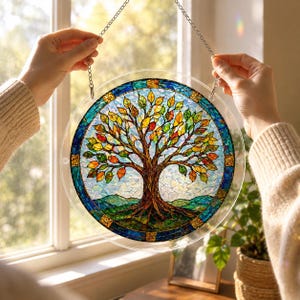 Pu&ograve; includere: Un acchiappasole circolare con un design dell'albero della vita in stile vetrata con foglie multicolori nelle tonalit&agrave; del rosso, arancione, giallo e verde. L'albero ha un tronco e radici marroni, su uno sfondo di cielo blu e colline verdi.