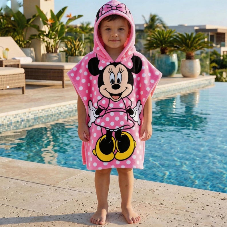 May include: A pink hooded towel featuring Minnie Mouse. The towel has a polka dot pattern and a hood with a bow. The Minnie Mouse graphic is centered on the front. The towel is worn by a child standing near a pool.
