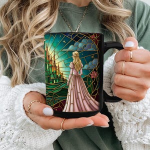 Puede incluir: Taza de cer&aacute;mica negra con un dise&ntilde;o de vidriera que representa a una mujer con un vestido rosa contemplando un castillo verde. La obra de arte incluye un cielo estrellado y detalles dorados. La taza tiene un asa negra.