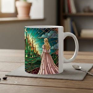 Puede incluir: Taza de cer&aacute;mica blanca con un dise&ntilde;o de vidriera. La obra de arte representa a una mujer con un vestido rosa mirando un castillo verde bajo un cielo estrellado. La taza tiene un asa curva y est&aacute; sobre un salvamanteles gris.