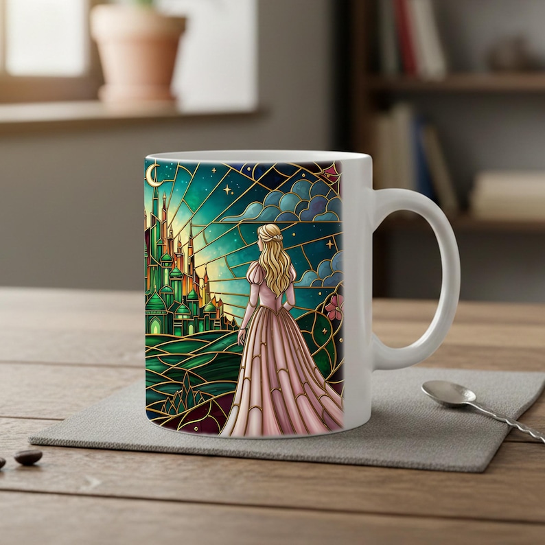Puede incluir: Taza de cer&aacute;mica blanca con un dise&ntilde;o de vidriera. La obra de arte representa a una mujer con un vestido rosa mirando un castillo verde bajo un cielo estrellado. La taza tiene un asa curva y est&aacute; sobre un salvamanteles gris.