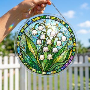 Pu&ograve; includere: Un acchiappasole circolare in vetro colorato raffigurante un grappolo di mughetti bianchi con foglie verdi. Il disegno &egrave; posto su uno sfondo testurizzato blu e verde iridescente, racchiuso da un bordo di piccole tessere di vetro.