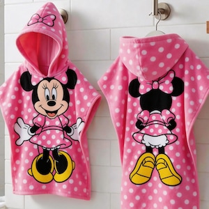May include: Pink hooded towel with white polka dots and a Minnie Mouse graphic. The towel features a hood with a bow and a large Minnie Mouse character on the front. The back of the towel also features a Minnie Mouse graphic.