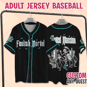 Puede incluir: Una camiseta de béisbol negra con botones y ribetes en color cerceta. El frente presenta el texto "Foolish Mortal" en blanco con murciélagos. La parte posterior muestra "Haunted Mansion" y tres personajes con sombreros de copa.