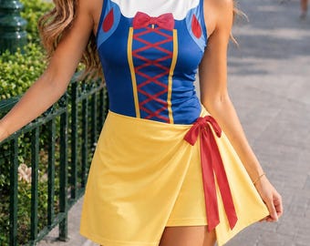 May include: A sleeveless bodysuit featuring a white upper bodice, a blue midsection with a red lace-up corset print, and a red bow detail. The garment includes a bright yellow wrap-style skirt overlay with a red ribbon tie at the side hip.