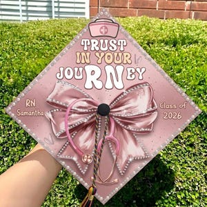 May include: A pink graduation cap decorated with a large bow, stethoscope, and the words "Trust in Your Journey RN." The cap is embellished with pearls and the text "Class of 2026" and "RN Samantha."