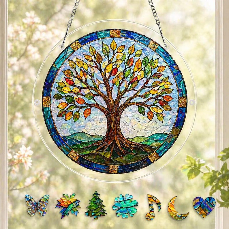 Pu&ograve; includere: Un acchiappasole circolare in vetro colorato raffigurante un albero con foglie multicolori, un tronco marrone e colline verdi, incorniciato da un bordo blu. Sotto ci sono sette piccoli ornamenti in vetro: farfalla, foglia, albero, fiore, nota musicale, luna e cuore.