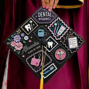 Può includere: Un cappello di laurea nero decorato con abbellimenti a tema dentale. Il cappello presenta le parole "Dental Hygienist" e "Alexandra Class of 2026". Gli abbellimenti includono denti, strumenti odontoiatrici, filo interdentale e un simbolo di caduceo.