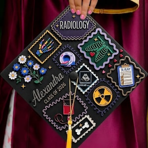 May include: Black graduation cap decorated with radiology-themed embellishments. Features the name "Alexandra" and "Class of 2026". Embellishments include a skeleton, flowers, and the word "Radiology".