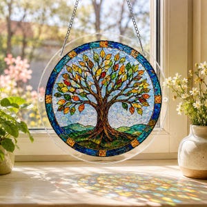 Pu&ograve; includere: Un acchiappasole circolare in vetro colorato raffigurante un albero con foglie multicolori nelle tonalit&agrave; del rosso, arancione, giallo e verde. L'albero &egrave; su uno sfondo azzurro con colline verdi alla base, racchiuso in un bordo blu.