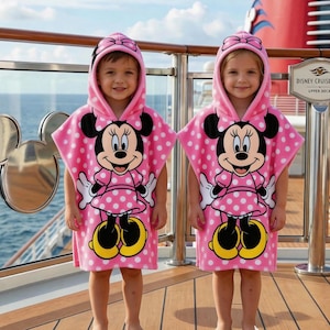 May include: Two children wearing pink hooded ponchos with white polka dots and a Minnie Mouse graphic. The ponchos have a hood with a bow and are made of a soft material. The children are standing on a wooden deck.