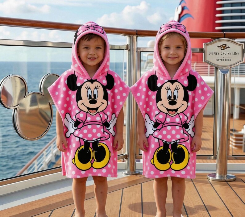 May include: Two children wearing pink hooded ponchos with white polka dots and a Minnie Mouse graphic. The ponchos have a hood with a bow and are made of a soft material. The children are standing on a wooden deck.