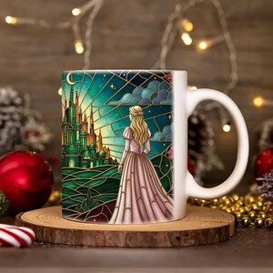 Puede incluir: Taza de cer&aacute;mica blanca con una ilustraci&oacute;n estilo vidriera de una mujer con un vestido rosa mirando un castillo verde bajo un cielo estrellado. La taza est&aacute; sobre un posavasos de madera, con decoraciones navide&ntilde;as en el fondo.