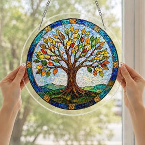 Pu&ograve; includere: Un acchiappasole circolare in vetro colorato raffigurante un albero con foglie multicolori nelle tonalit&agrave; del giallo, arancione e verde. Il tronco &egrave; marrone, su uno sfondo di cielo azzurro e colline verdi, racchiuso da un bordo blu.