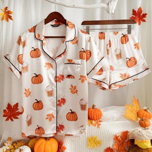 Peut inclure: Ensemble de pyjama en satin blanc avec imprimé citrouilles et feuilles d'automne. Le haut à manches courtes et le short ont une bordure noire. L'ensemble comprend une chemise boutonnée avec une poche poitrine et un short assorti.