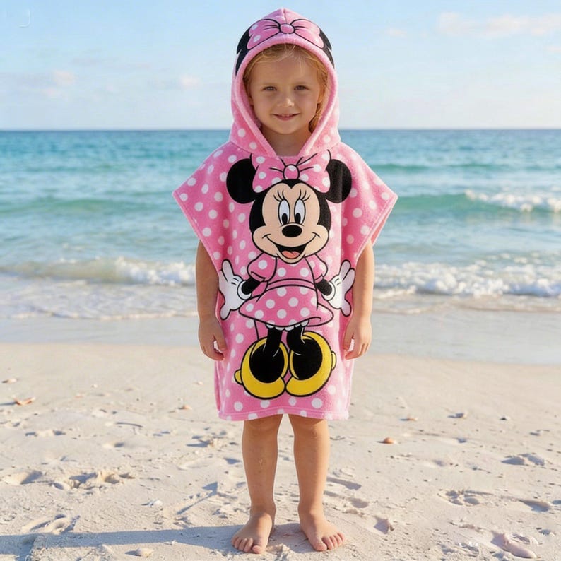 May include: A pink hooded beach towel featuring Minnie Mouse. The towel has a polka dot pattern and a hood with a bow. Minnie Mouse is depicted in the center of the towel. The child is standing on a sandy beach with the ocean in the background.