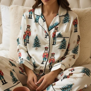 Puede incluir: Conjunto de pijama blanco con temática navideña. El conjunto presenta una camisa con botones y pantalones a juego, ambos adornados con un estampado de cascanueces, árboles de Navidad y copos de nieve. Ribete negro en el cuello, puños y bolsillos.