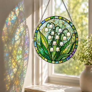 Pu&ograve; includere: Un acchiappasole circolare in vetro colorato raffigurante un grappolo di mughetti bianchi con foglie verdi, racchiuso da un bordo decorativo di pezzi di vetro blu, verdi e gialli, sospeso tramite una sottile catena metallica davanti a una finestra.