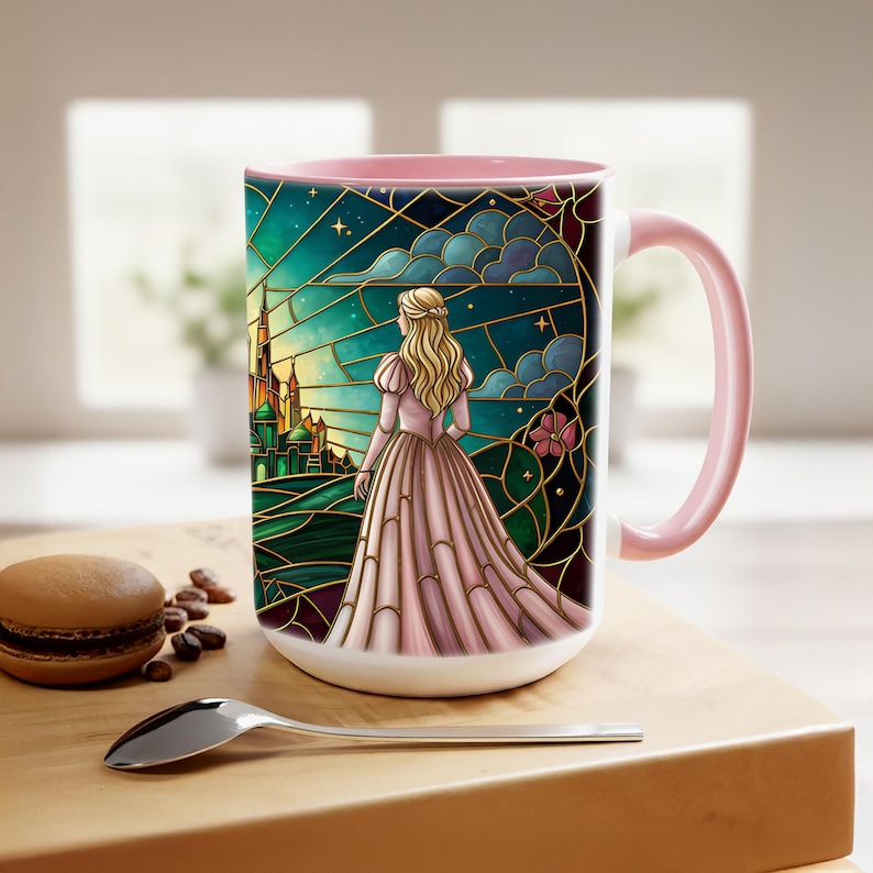 Puede incluir: Taza de cer&aacute;mica blanca con interior y asa rosas. La taza presenta una ilustraci&oacute;n estilo vidriera de una mujer con un vestido rosa, frente a un castillo. El fondo es una mezcla de verde, azul y dorado.