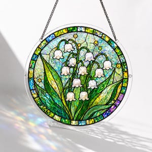 Pu&ograve; includere: Un acchiappasole circolare in vetro colorato raffigurante un grappolo di mughetti bianchi con foglie verdi. Il design &egrave; posto su uno sfondo testurizzato e iridescente con un bordo di piccoli pezzi di vetro verdi, blu e gialli.