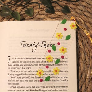 Op de afbeelding: Een bladwijzer met een gele kwast en een witte achtergrond versierd met gele bloemen, groene bladeren en rode stippen. De bladwijzer ligt op een boek met de zichtbare tekst "Twenty-Three".