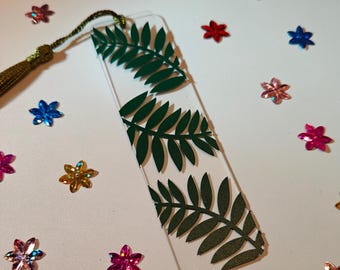 May include: A clear acrylic bookmark with a green fern design. The bookmark has a tassel attached to the top.